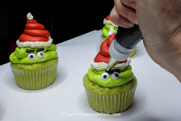 Green Grinch cupcakes with eyes and eyebrows being piped on with black icing