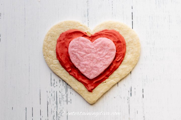 A smaller pink heart cookie stuck to a larger white heart cookie with red royal icing drawn in a heart design