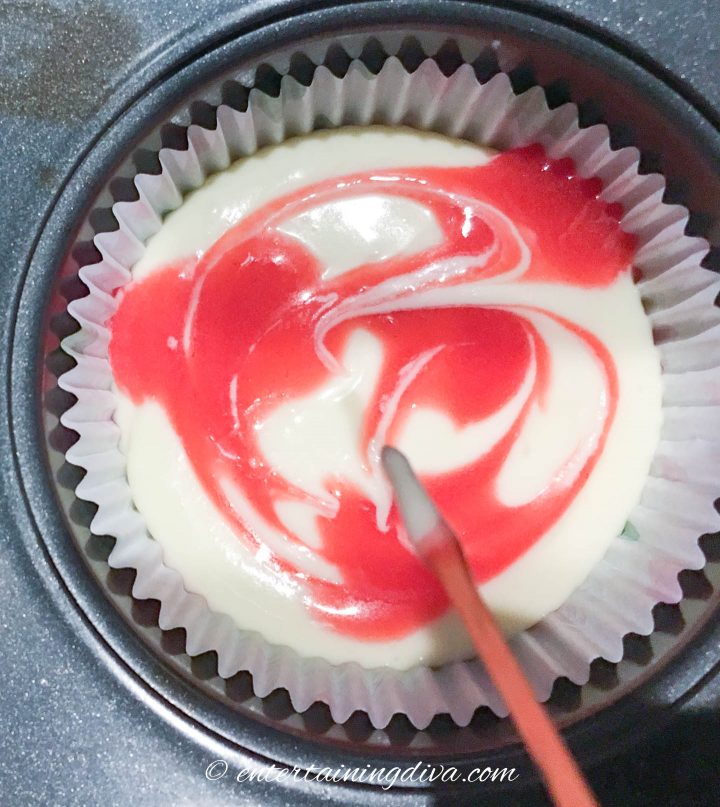 Raspberry sauce being swirled into the top of a mini cheesecake with a toothpick