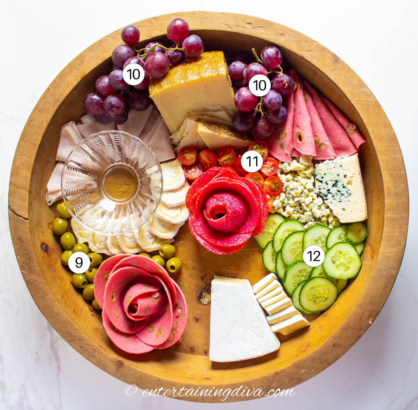 A round charcuterie board with cheeses, meats, olives, grapes, cherry tomatoes and cucumbers on it.