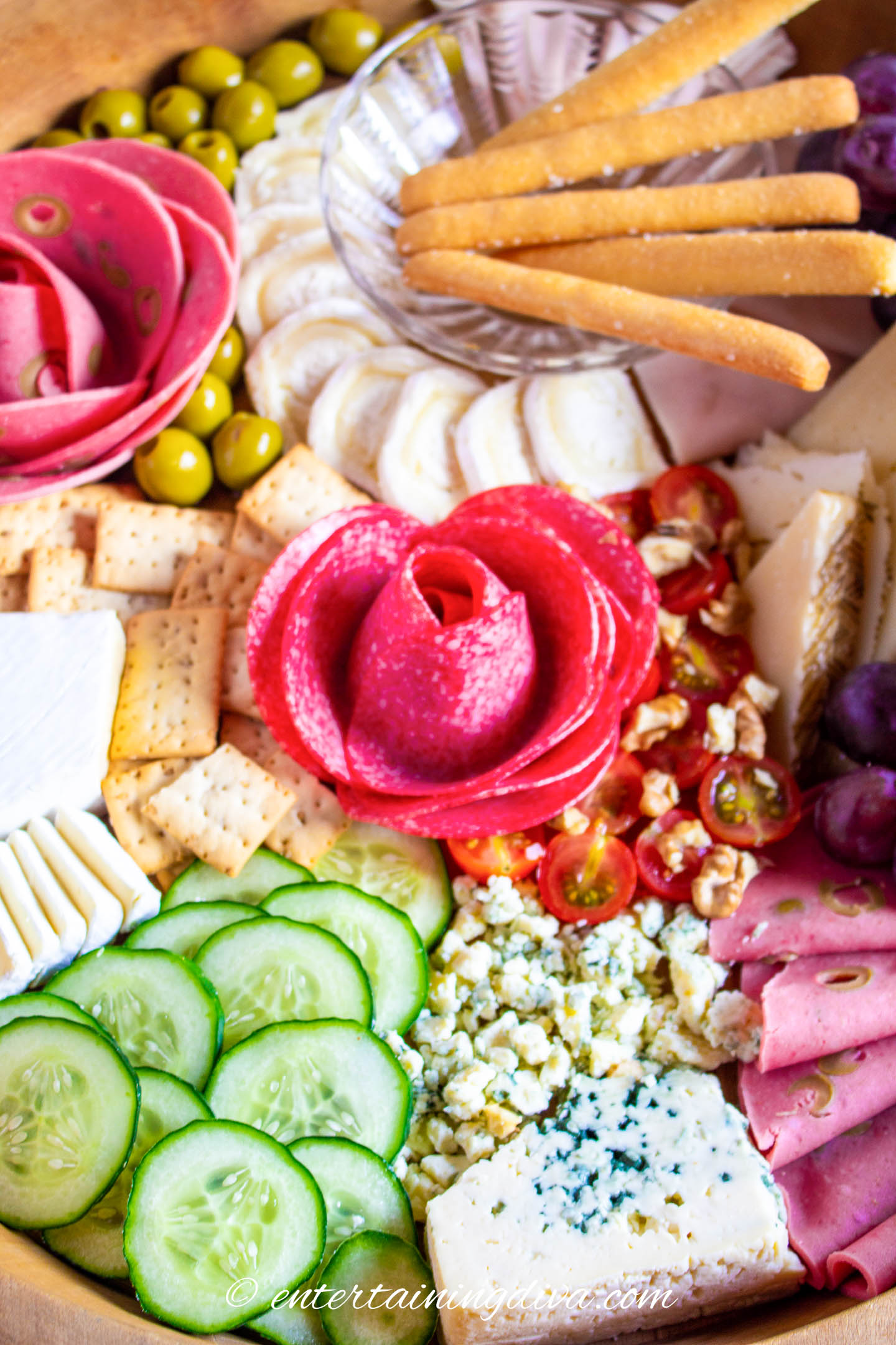 A charcuterie board with sliced meat in a flower shape, cucumbers, crackers breadsticks, cherry tomatoes, olives and gorgonzola cheese