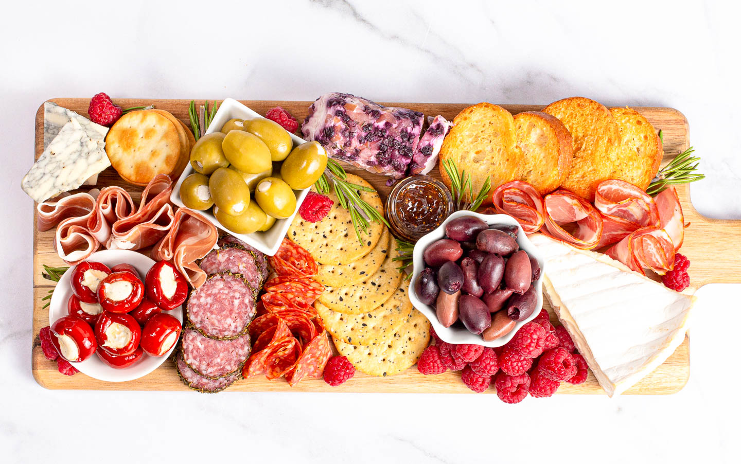 Four bowls filled with olives, stuffed peppers and jam on a long charcuterie board filled with meats, cheeses, raspberries and crackers