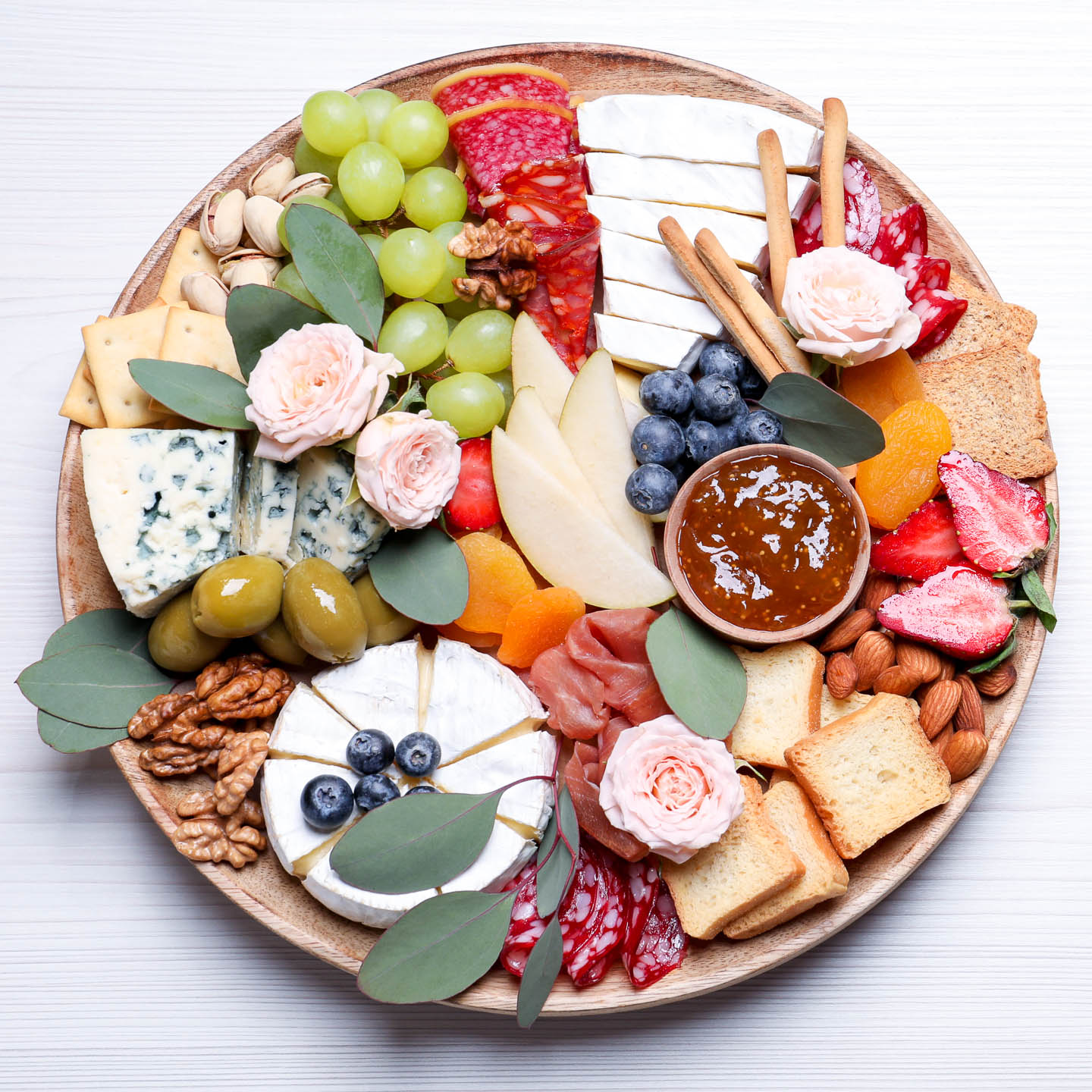 A round charcuterie board with cheeses, meats, jam, blueberries, grapes, apple slices, olives, apricots, nuts and roses on it