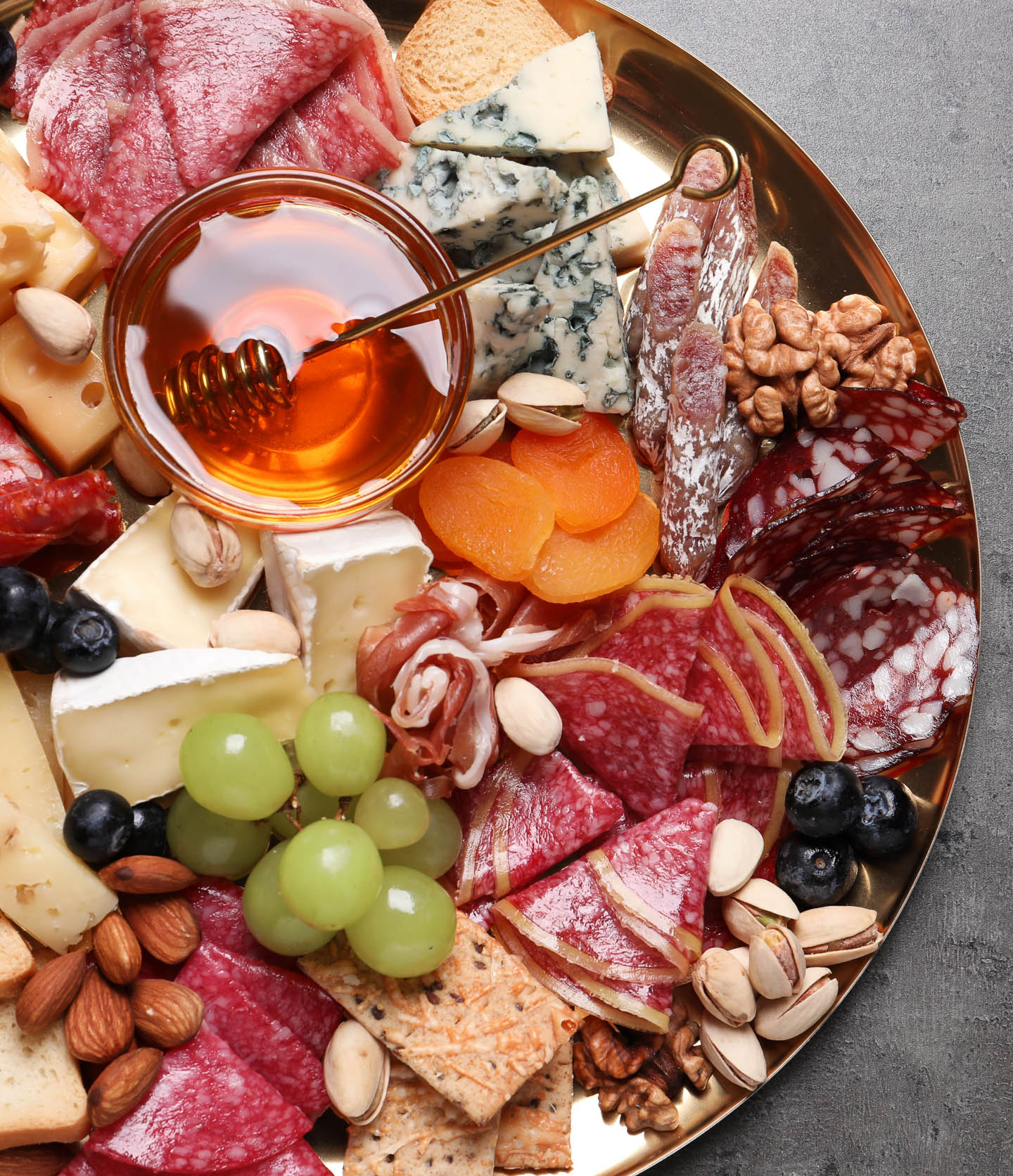 A round charcuterie board with meats, cheese, grapes, blubeberries, nuts, apricots and a bowl of honey.