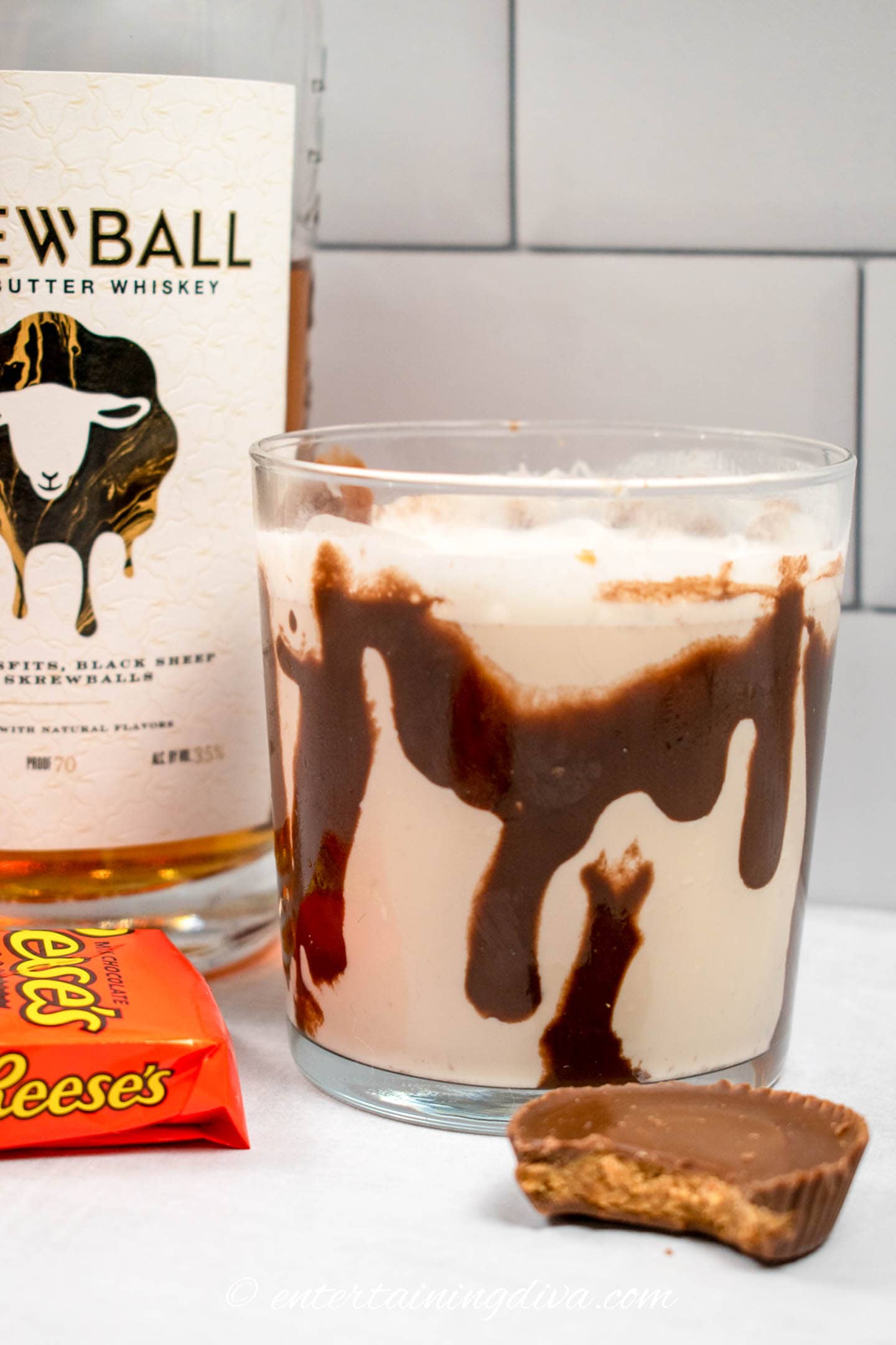 peanut butter whisky cocktail in a rocks glass drizzled with chocolate, beside a bottle of skrewball whiskey and a Reese's peanut butter cup