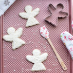 Decorated Christmas angel rolled sugar cookies on a baking sheet with an angel cookie cutter and a spatula
