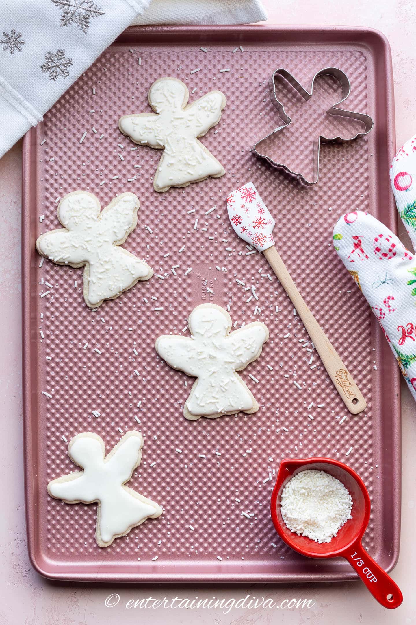 Decorated Christmas angel sugar cookies on a baking sheet with an angel cookie cutter, a spatula and a measuring cup