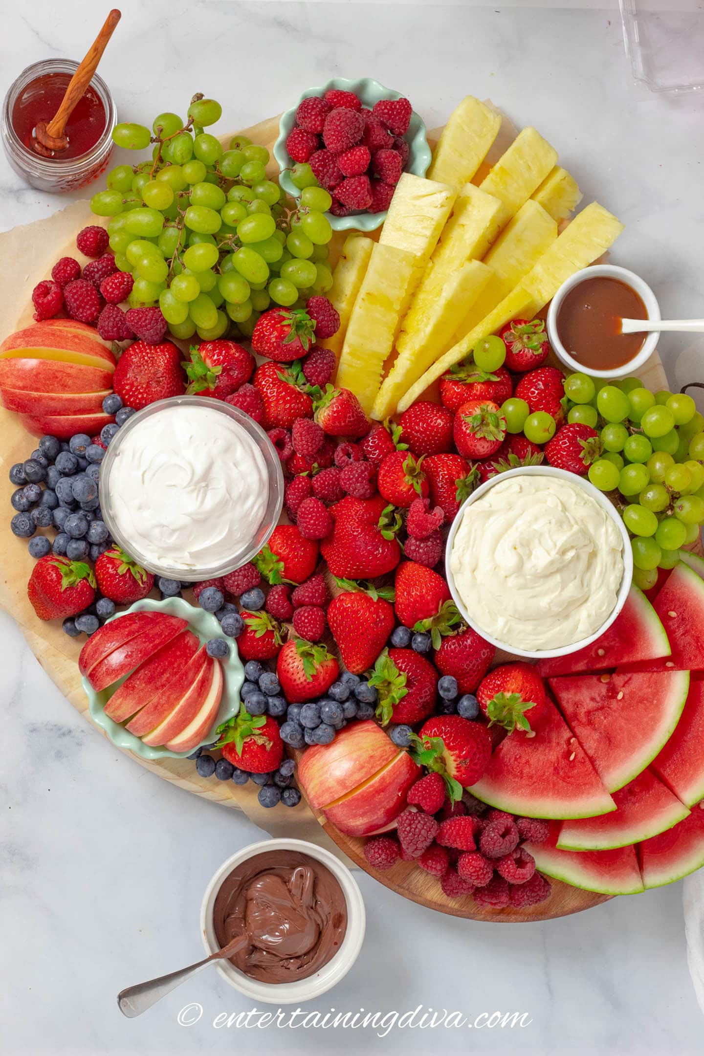 fruit charcuterie board with strawberries, raspberries, blueberries, apples, pineapple, watermelon, grapes and dips (whipped cream, cream cheese fruit dip, caramel, honey and hazel nut butter)