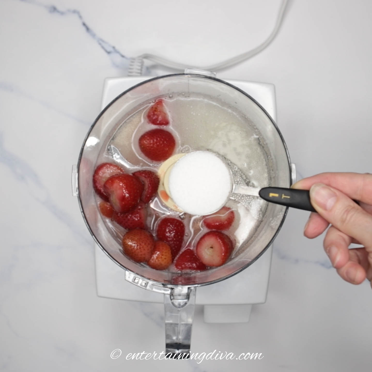 Sugar in a tablespoon above a food processor with strawberries and sparkling wine in it