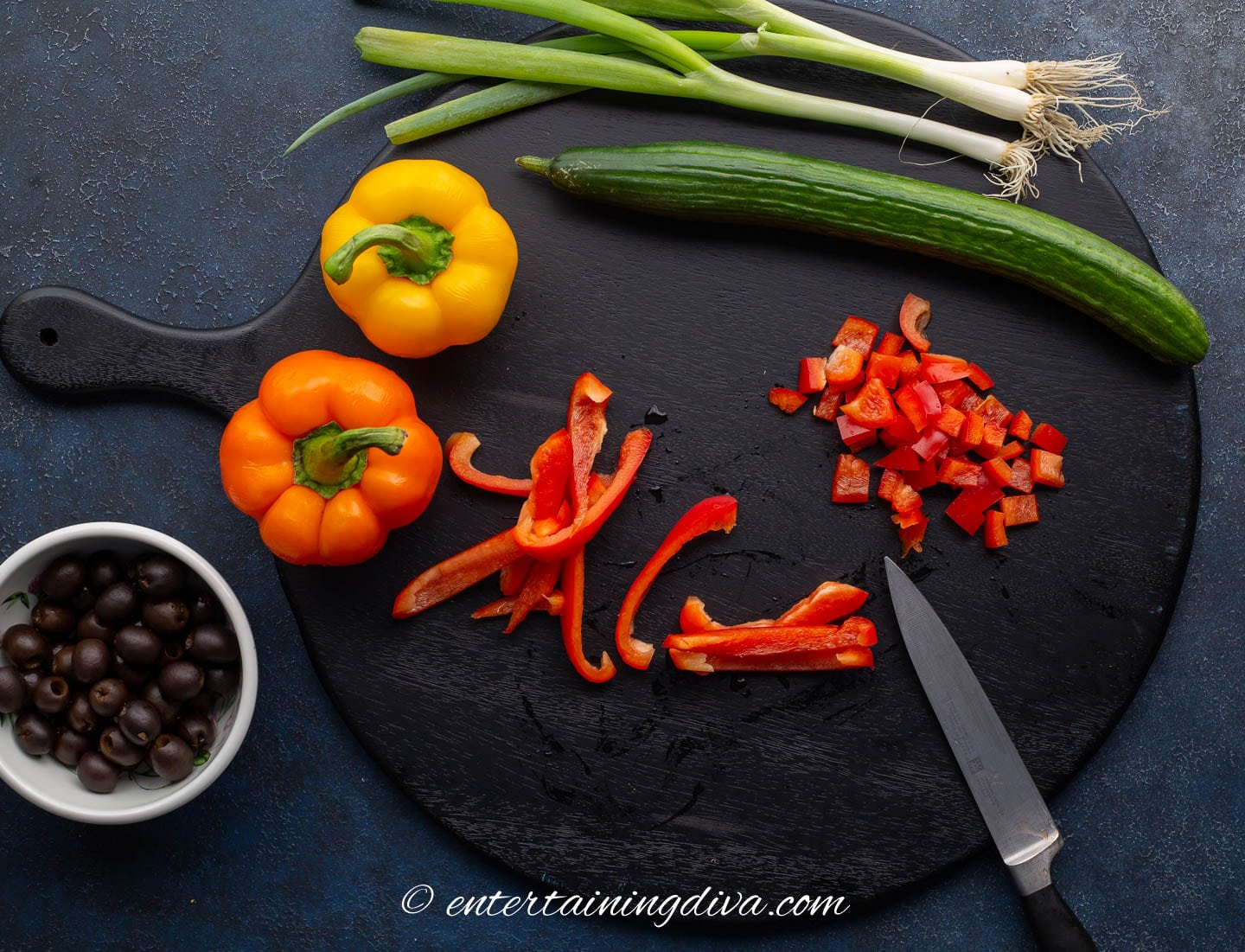Chopped red peppers on a cutting board with orange and yellow peppers, a cucumber, green onions and black olives