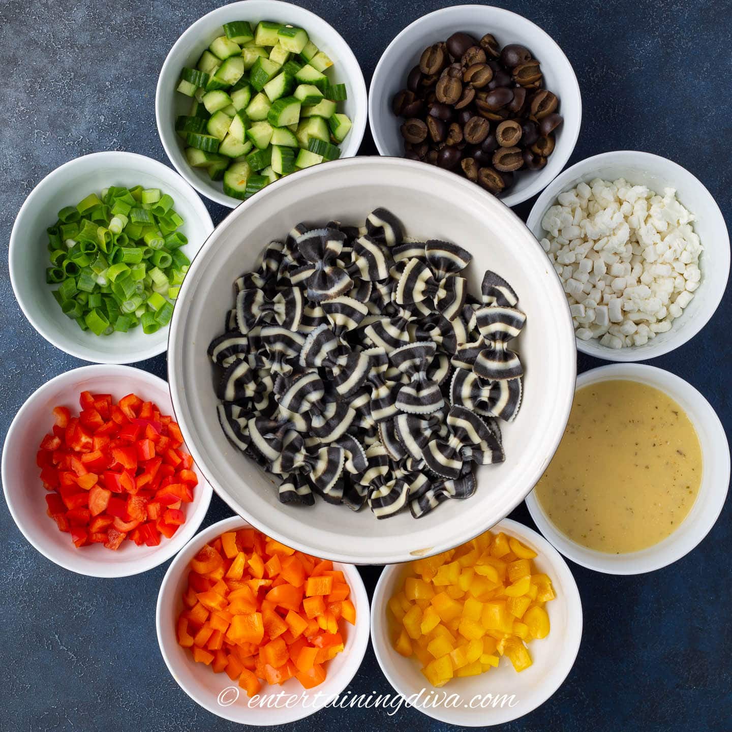 zebra bow tie pasta in a bowl surrounded by chopped cucumbers, black olives, goat cheese, Italian dressing, yellow peppers, orange peppers, red peppers and green onions in small bowls