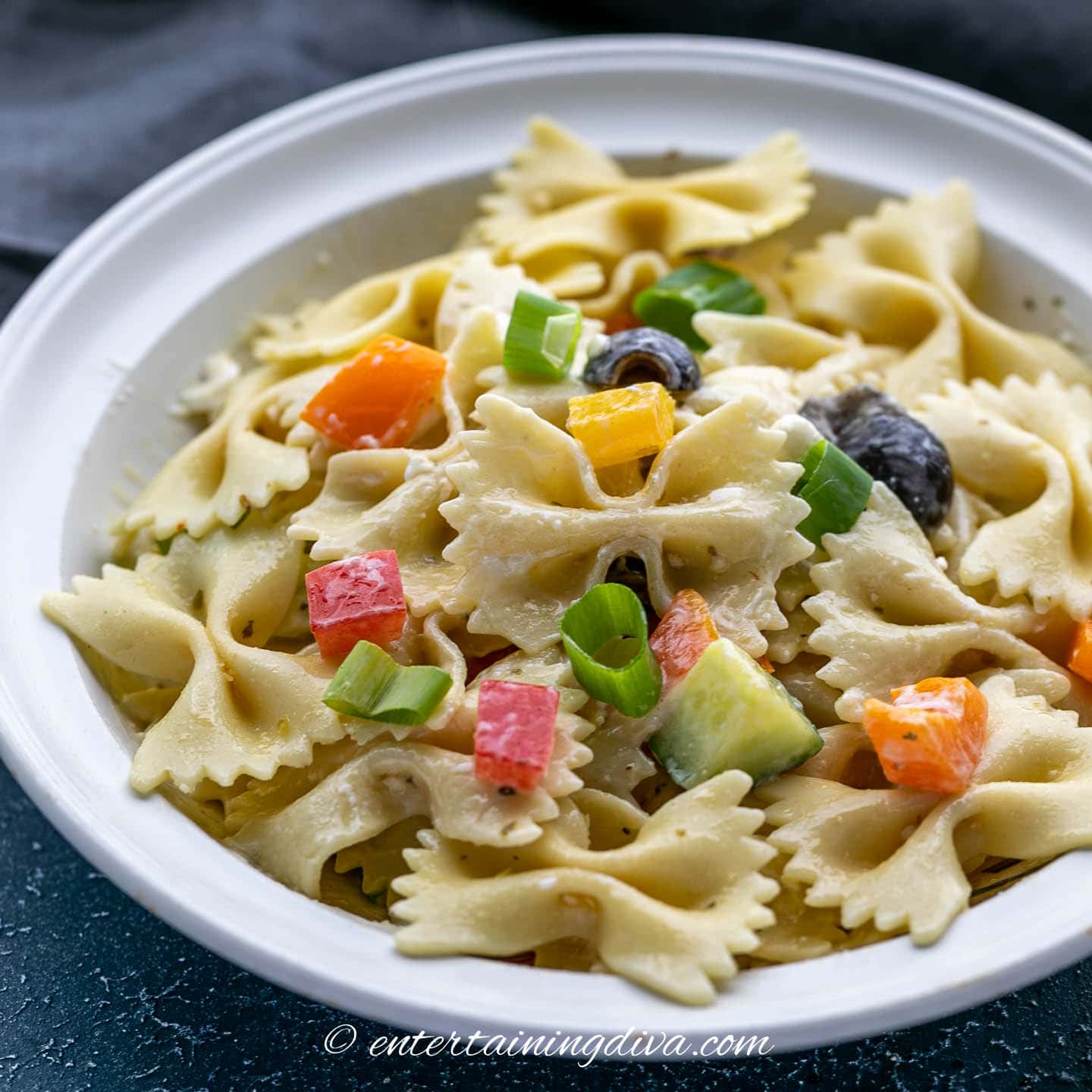 bow tie pasta salad with orange bell peppers, black olives, green onions, cucumbers and Italian dressing in a bowl