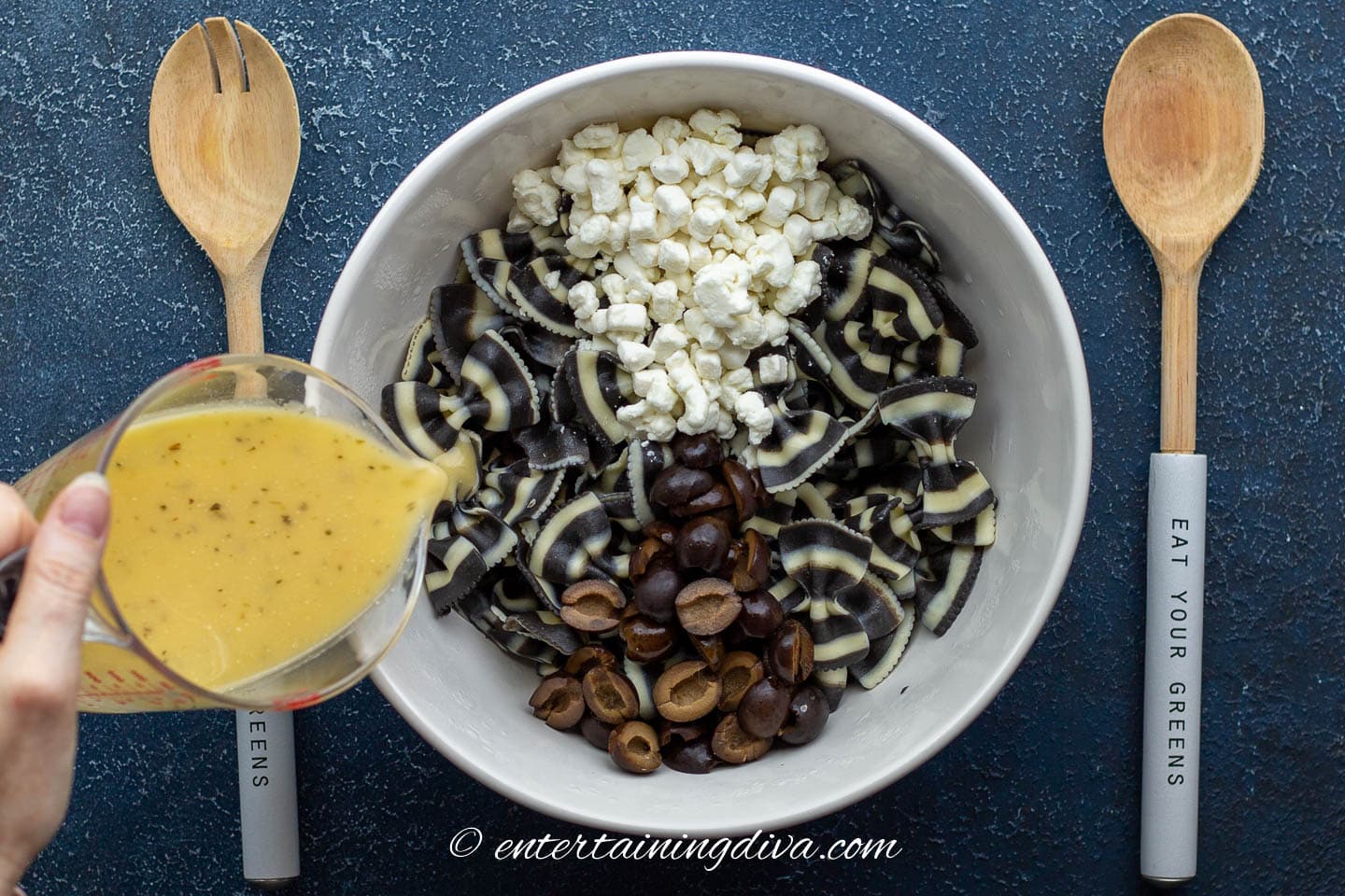 Italian dressing being poured over black and white bow tie pasta in a bowl with goat cheese and black olives
