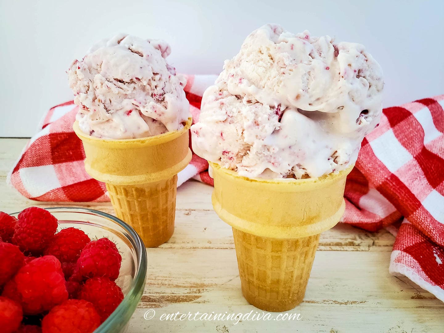 two raspberry ice cream cones with a bowl of raspberries in front or them and a red and white towel behind
