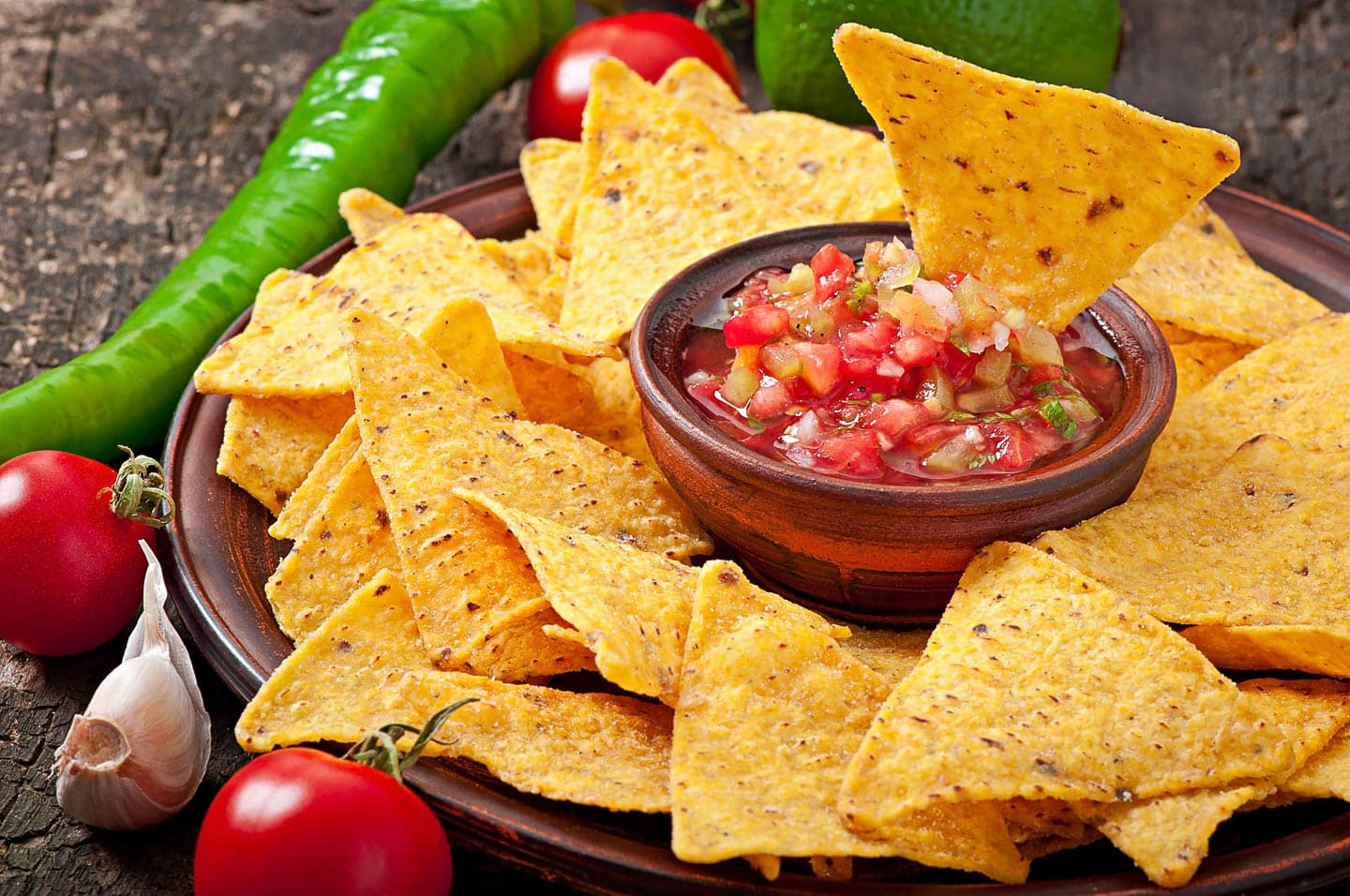 chip dipped in a bowl of Pico de gallo that is surrounded by chips, tomatoes, garlic and chile peppers.