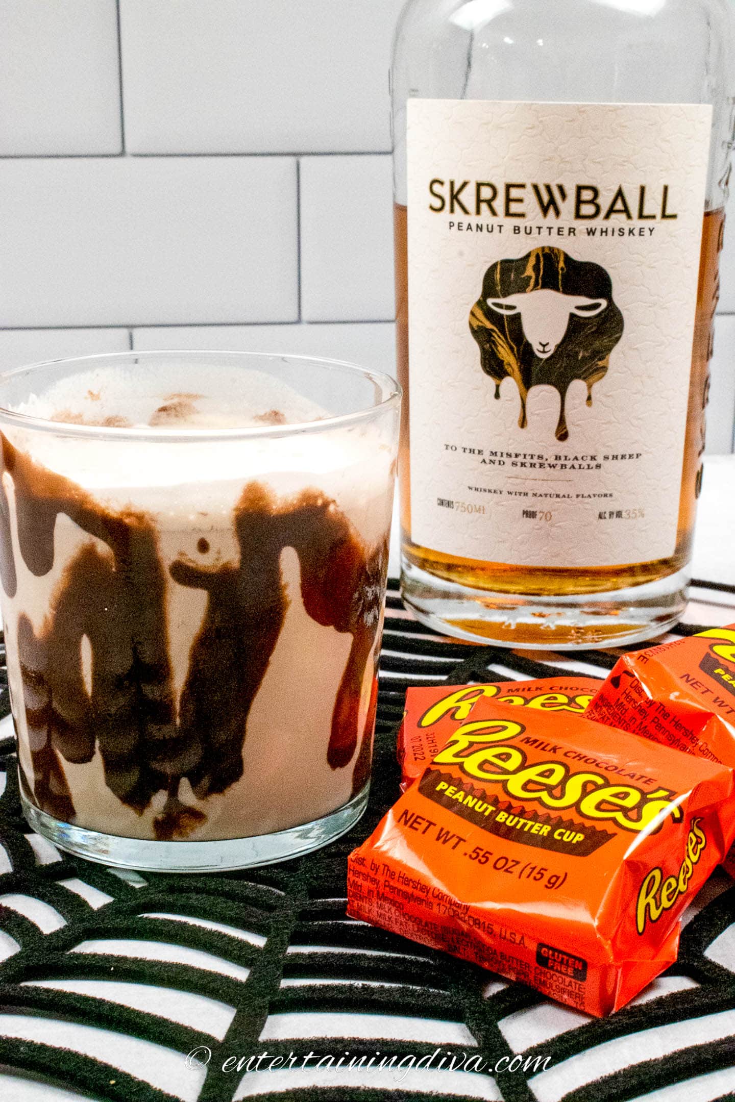 Peanut butter cup cocktail on a black spider web beside some Reese's peanut butter cups and a bottle of Skrewball whiskey