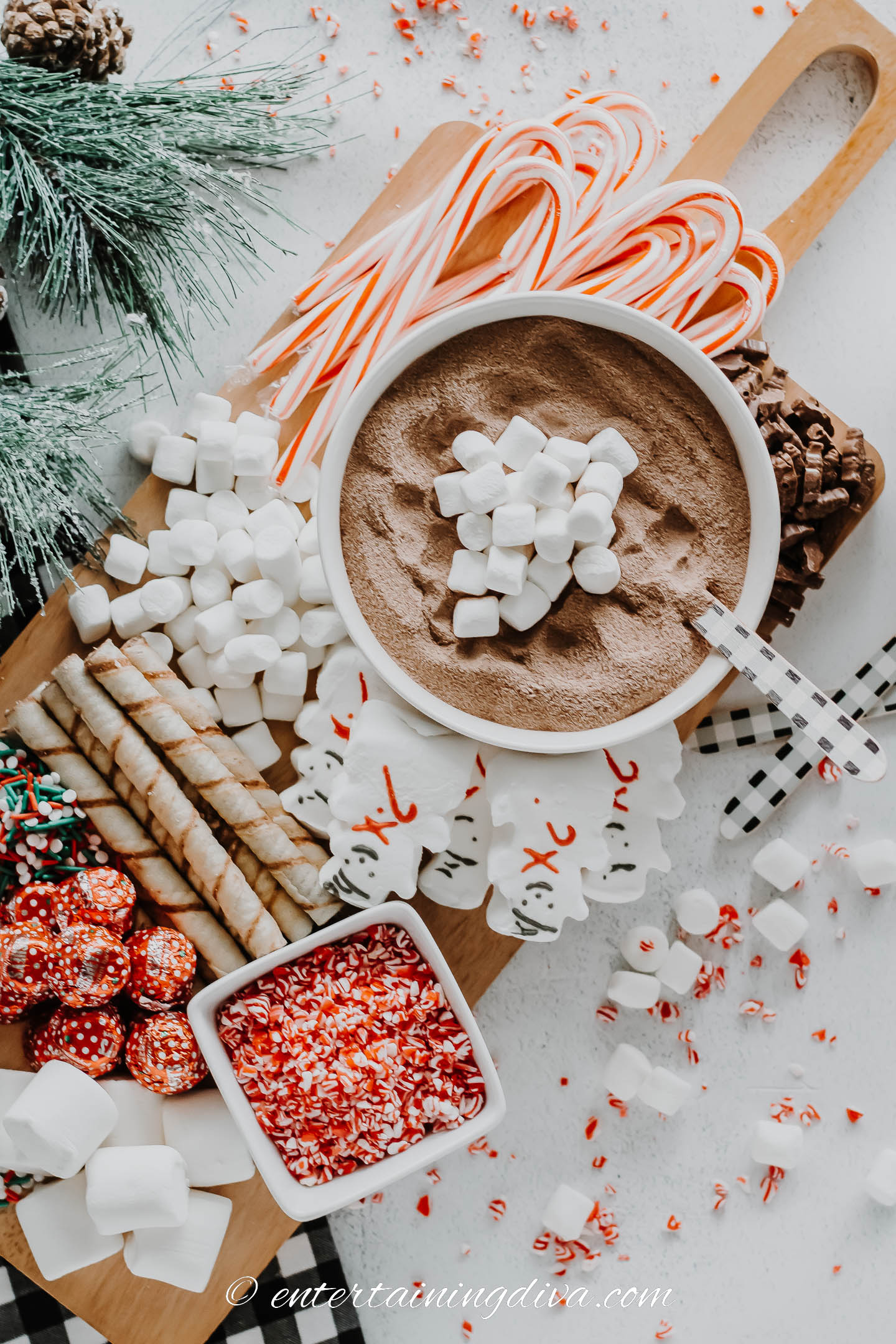 overhead of a hot chocolate charcuterie board with hot cocoa mix, mini marshmallows, candy canes, chocolate, snowman peeps, wafer cookies, and Christmas sprinkles