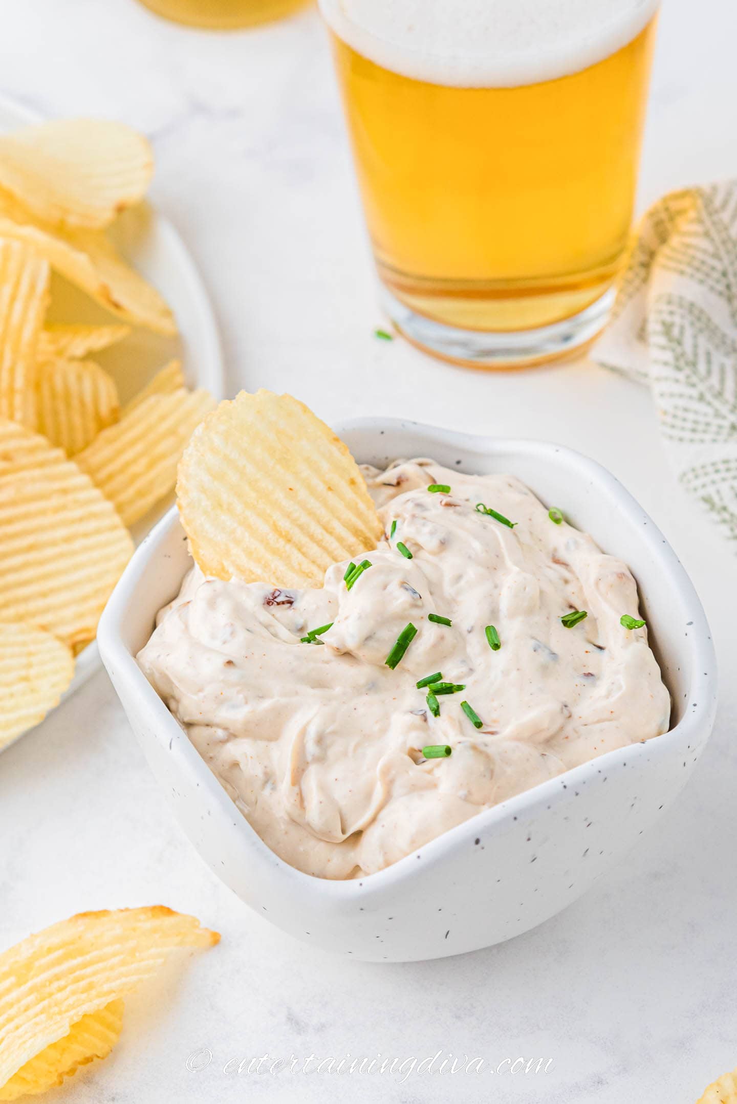 A bowl of caramelized onion dip in front of a plate of chips and a glass