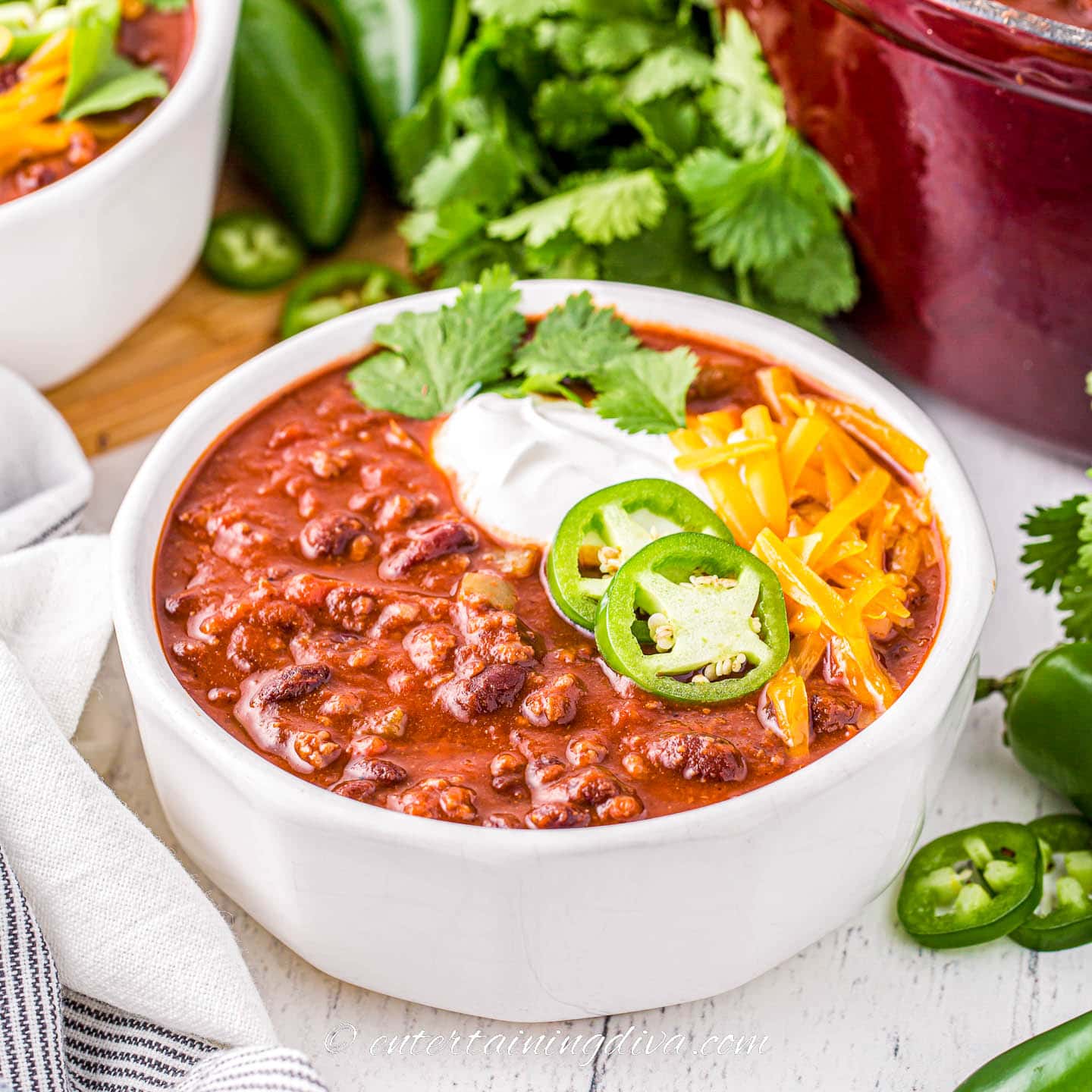 thick chili in a white bowl topped with jalapeno peppers, sour cream and grated cheese