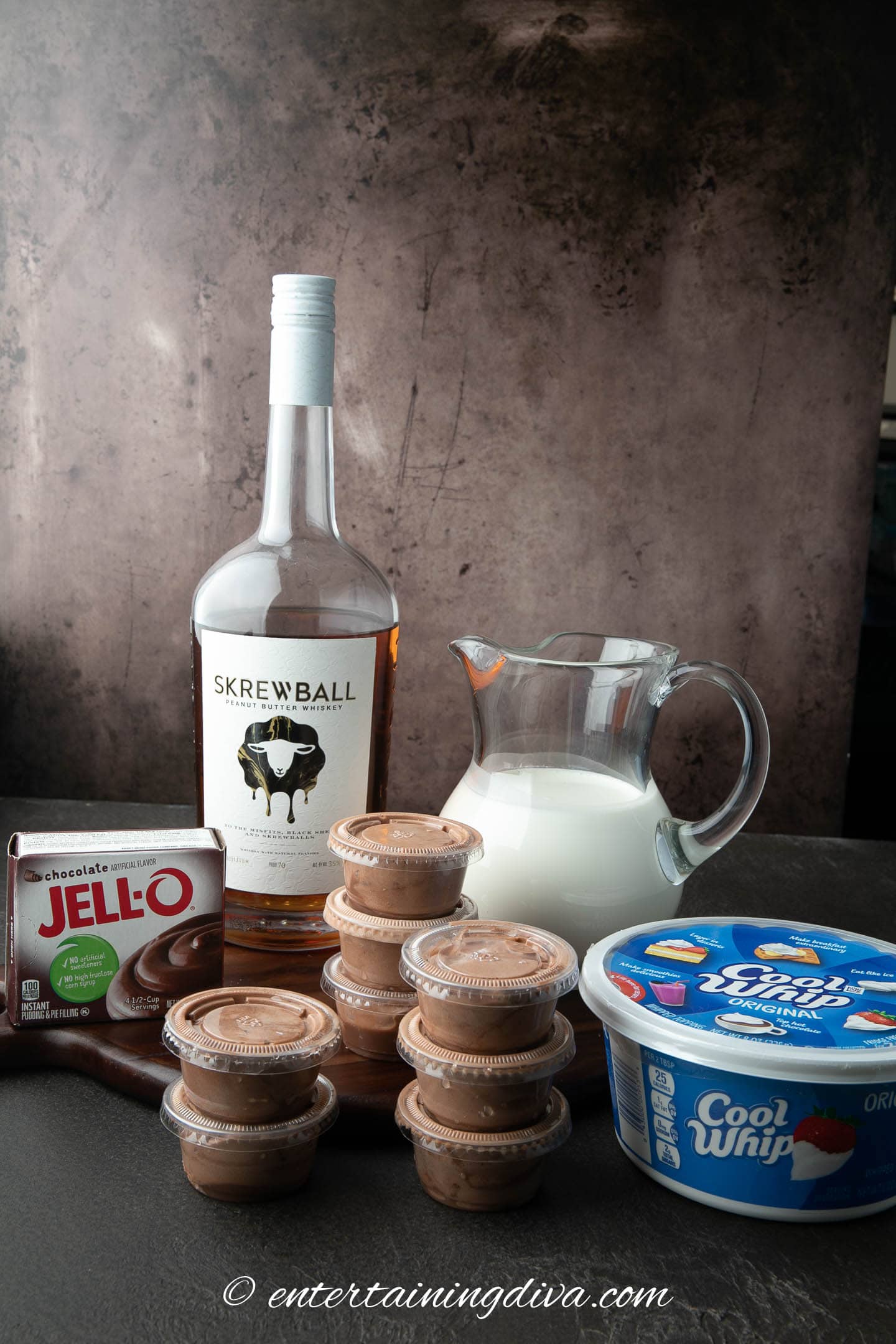 Peanut butter chocolate pudding shots with a box of chocolate pudding mix, a bottle of Skrewball peanut butter whiskey, a pitcher of milk and a container of whipped topping