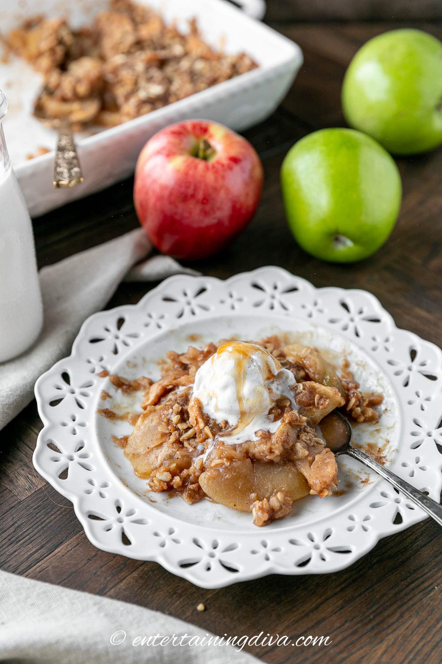 Apple oat crisp topped with vanilla ice cream and caramel sauce in front of some apples and a baking dish of apple crisp