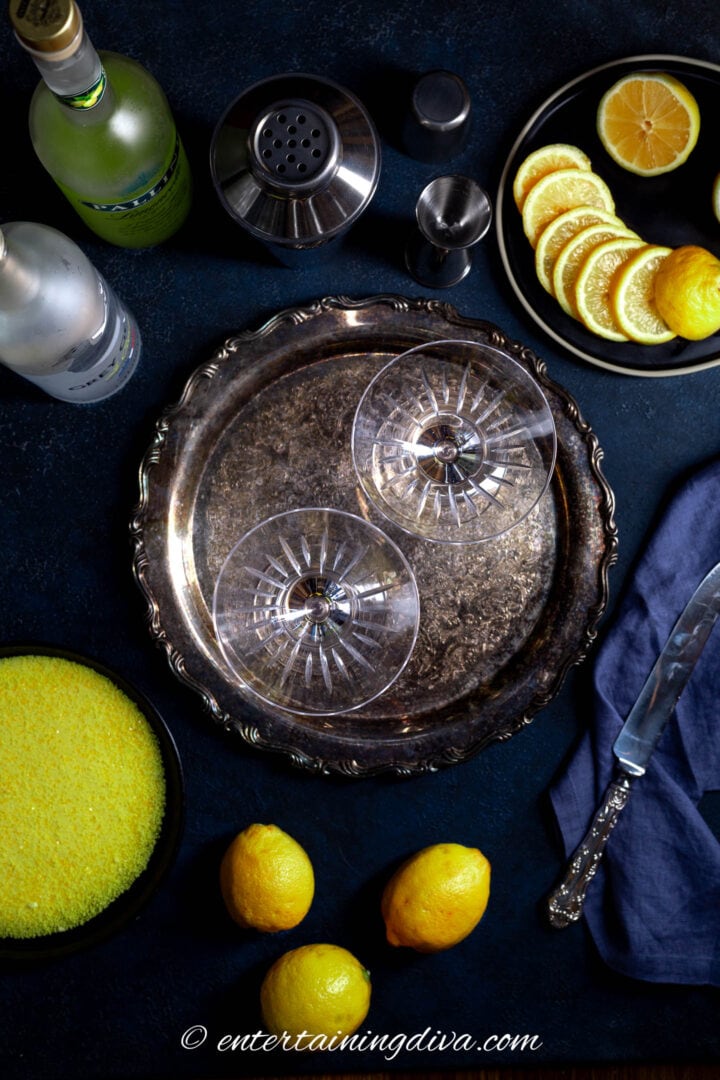A silver tray with martini glasses on it, beside lemons, rimming sugar and bottles of vodka and limoncello