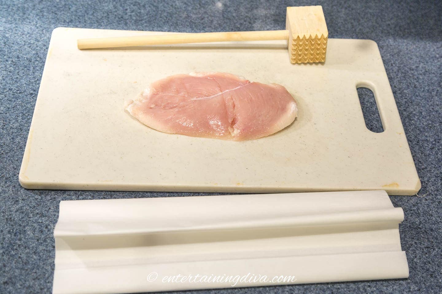 chicken breast on a cutting board with a meat tenderizer and parchment paper