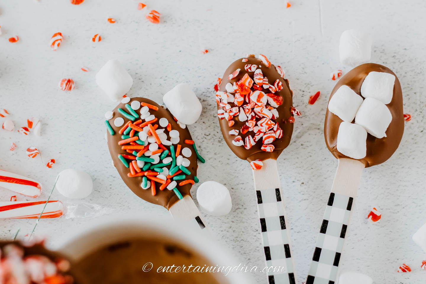 Three homemade hot chocolate stirring spoons - one with sprinkles, one with crushed candy canes and one with mini marshmallows