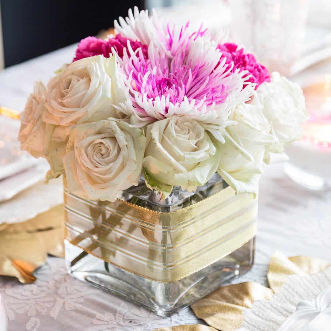 Pink and white centerpiece made with flowers and a square vase wrapped in gold ribbon