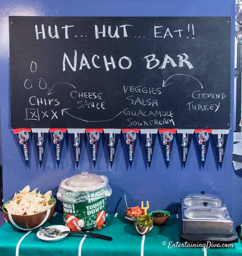 A help-yourself nacho bar with a bowl of nachos, a crockpot with cheese sauce, nacho toppings and ground beef under a blackboard decorated with Nacho Bar text