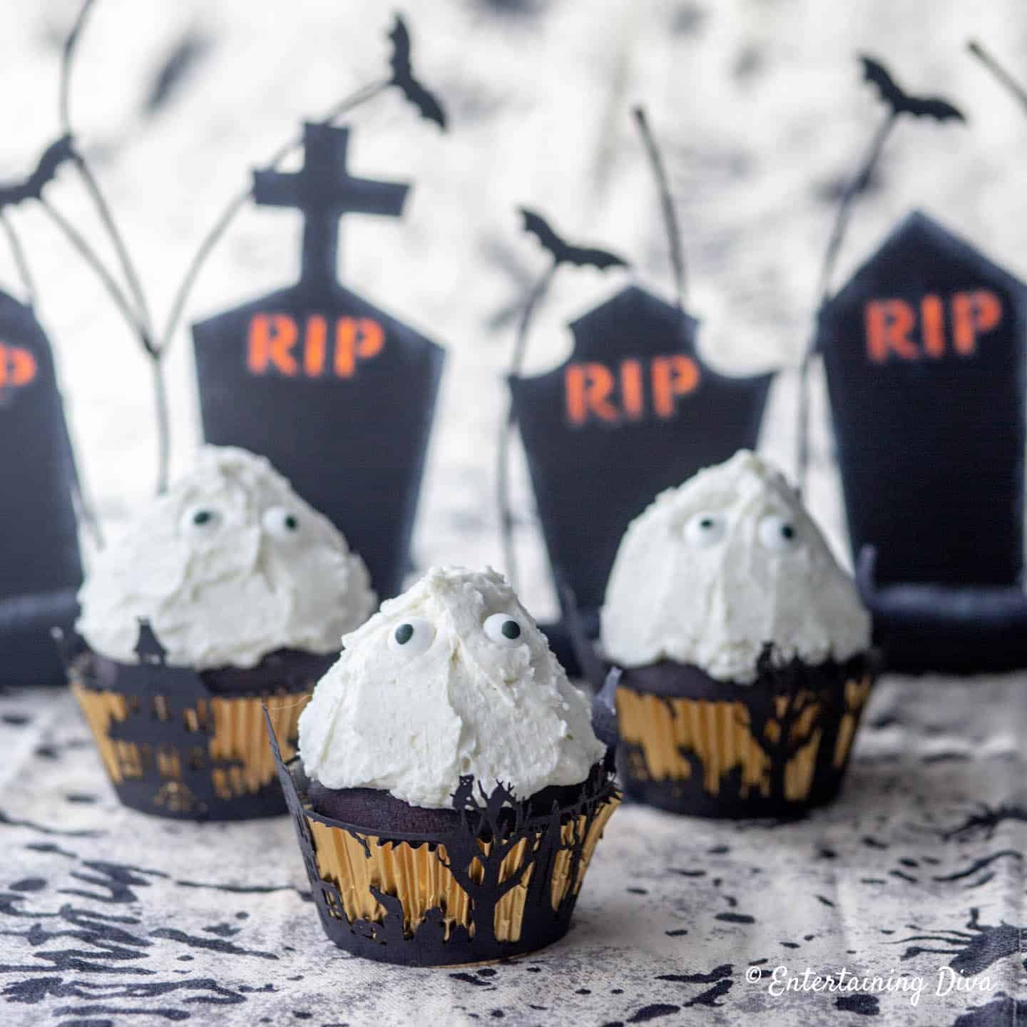 Chocolate cupcakes with white frosting and candy eyes are placed in black wrappers, with blurred “RIP” tombstone decorations in the background.