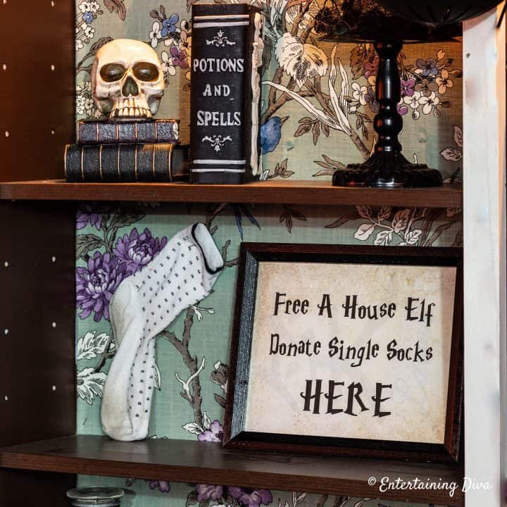 Framed printable sign that says "Free a house elf donate single socks HERE" displayed on a shelf with other Harry Potter party decorations