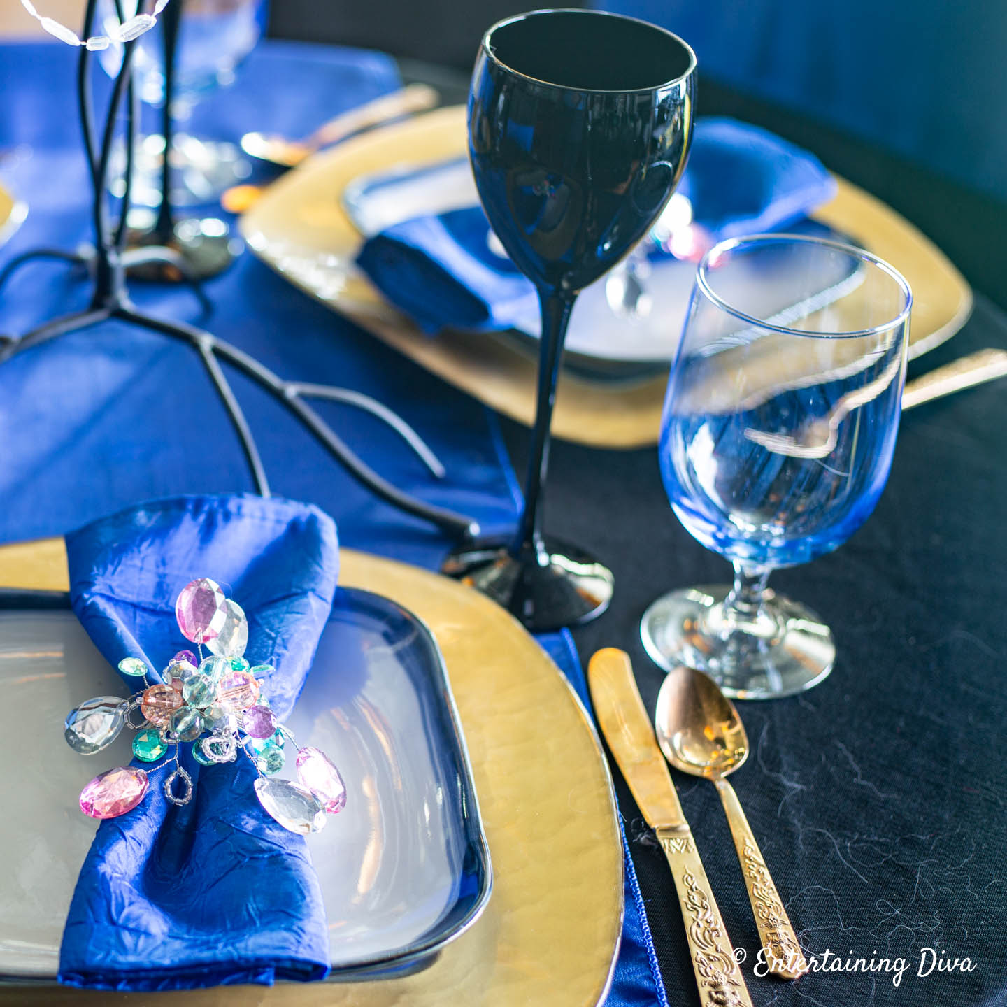 Place setting with a blue napkin and water glass, gold charger and cutlery, and black wine glasses