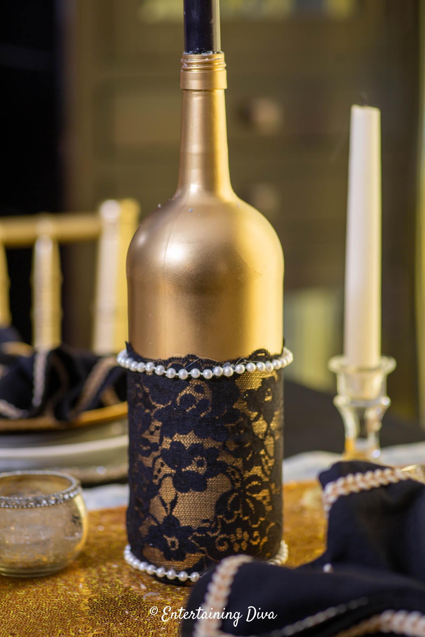 A gold wine bottle decorated with black lace and faux pearls