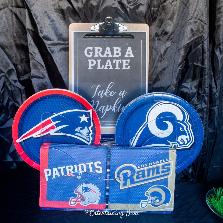 "Grab a plate" sign on a clipboard in front of Patriots and Rams plates and napkins