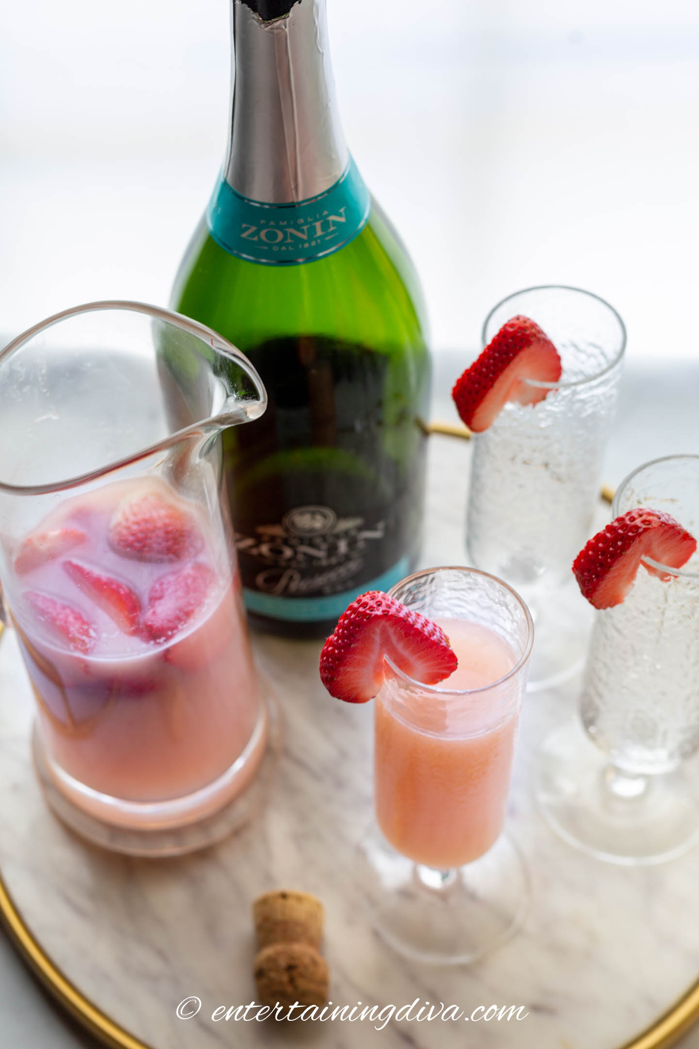 A pitcher of strawberry mimosas with 3 glasses and a bottle of Prosecco