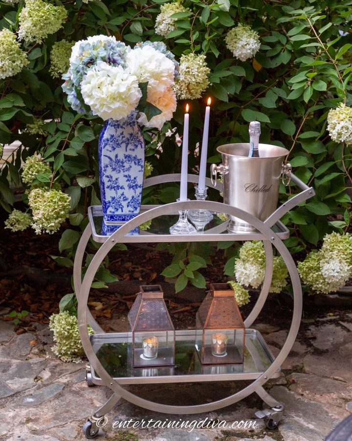 Bar cart with flowers, candles and champagne on a patio in front of a hydrangea