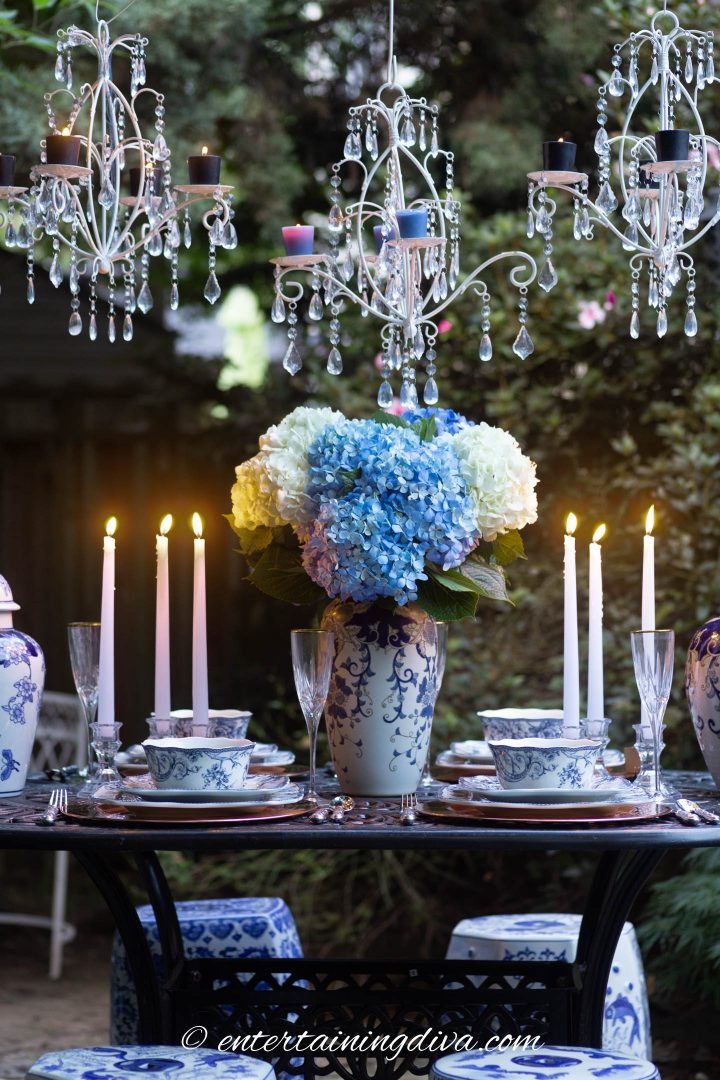Three white candle chandeliers hung over a blue and white outdoor dinner party table setting