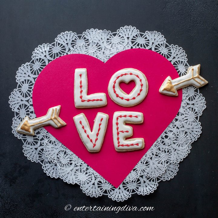 Valentine sugar cookies in a LOVE shape with an arrow on either side and displayed on a red paper heart with a white paper doily