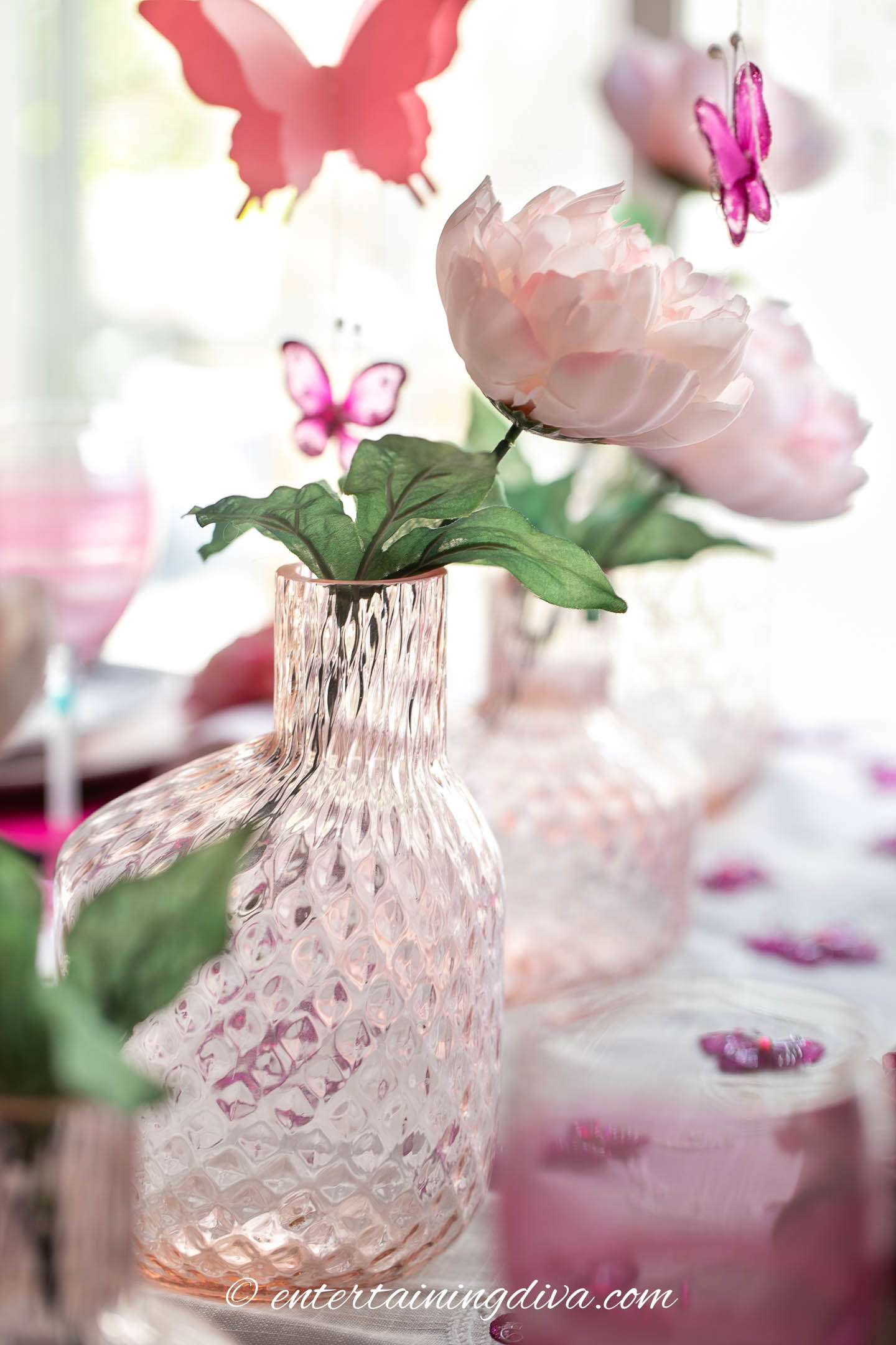 Table centerpiece with pink flowers in pink glass vases and butterflies hanging above them