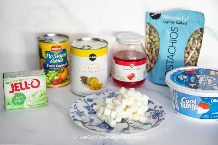pistachio Jell-O, fruit cocktail, pineapple, marshmallows, maraschino cherries, pistachios, and Cool Whip on white surface