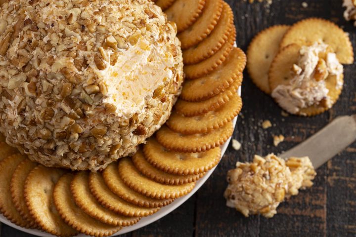 Top view of the cheeseball with a chunk taken out of it on a plate of crackers beside a knife with the cheese mixture on it