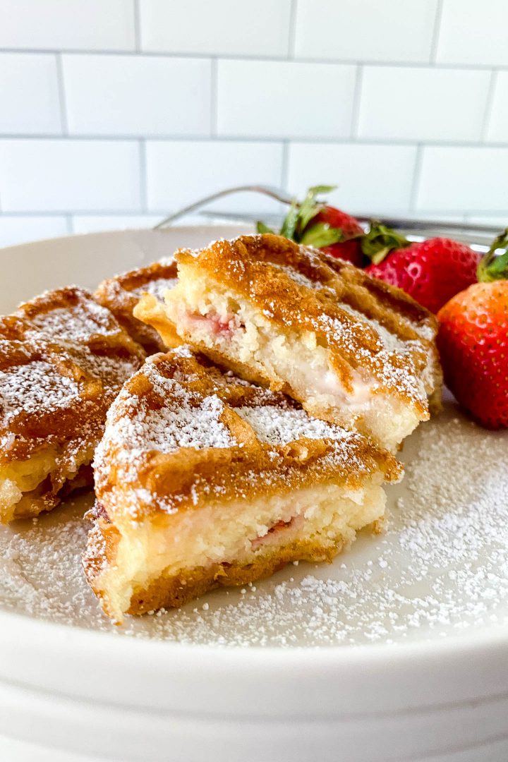 Strawberry cheesecake stuffed waffles sprinkled with icing sugar and served with strawberries on a white plate