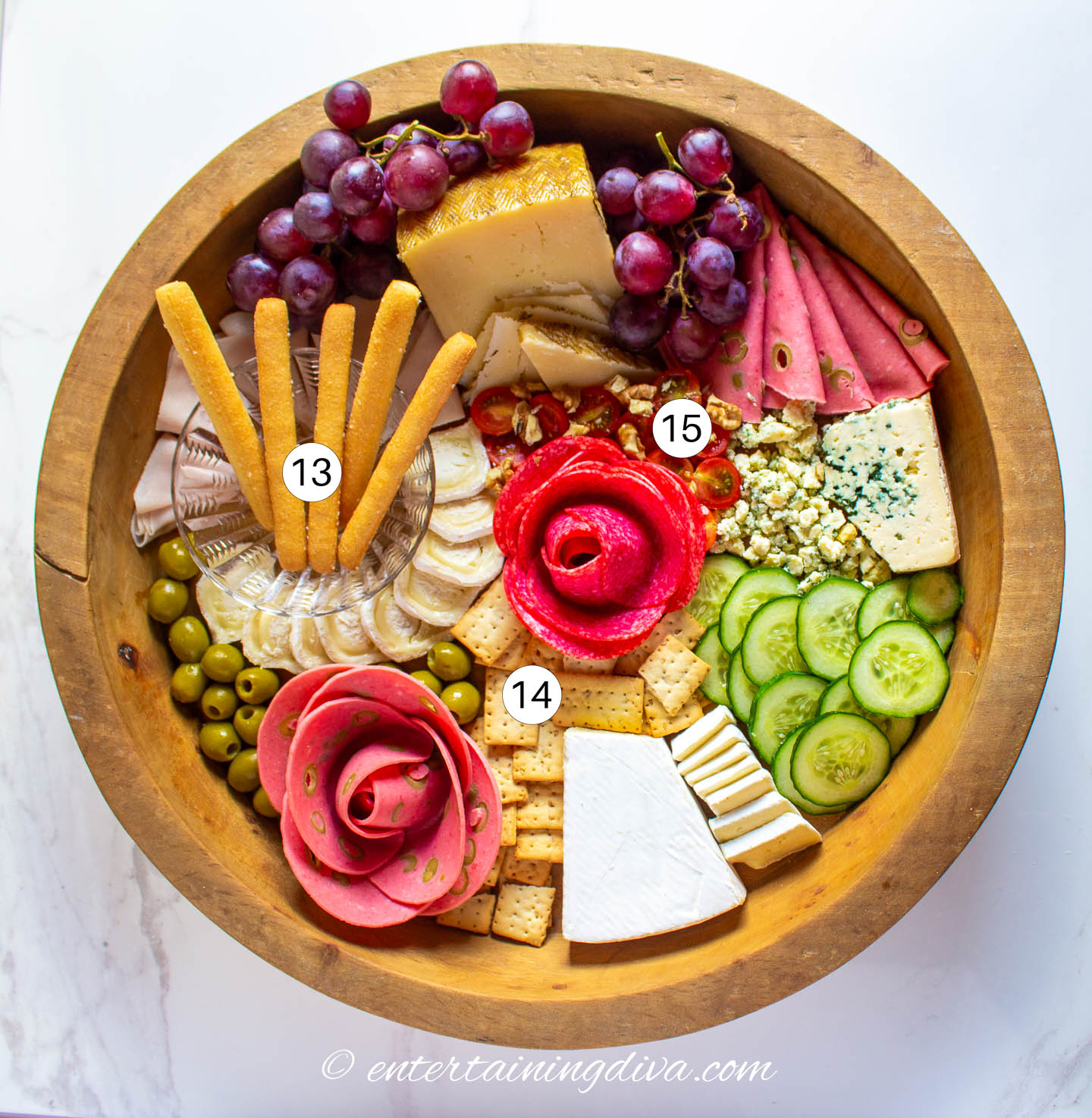 A round charcuterie board with meats, cheeses, fruits, vegetables, breadsticks, crackers and walnuts arranged on it