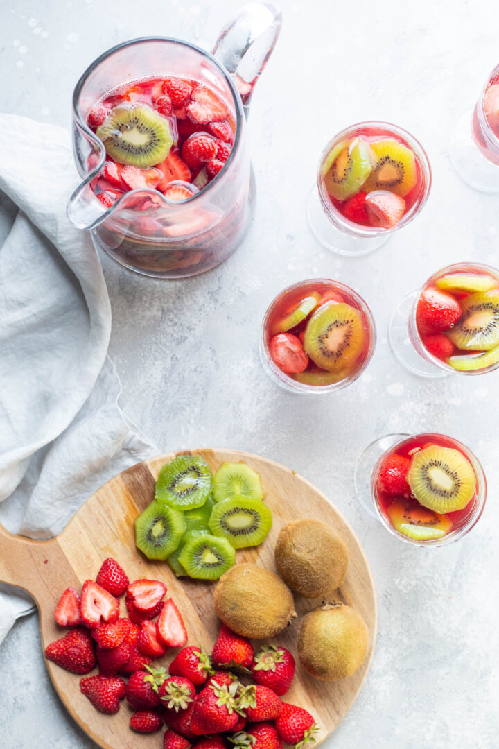 Overhead of strawberry kiwi sangria in a pitcher with a few glasses and a cutting board with kiwis and strawberries on it
