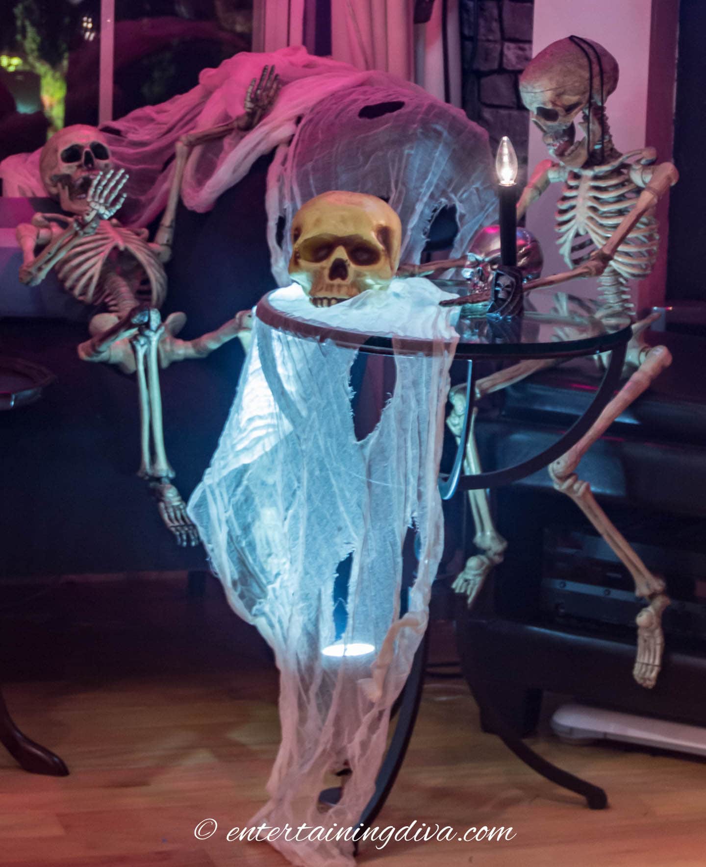 Two skeletons sitting around a small glass table with a skull, a candle and some white creepy cloth that is lit from below