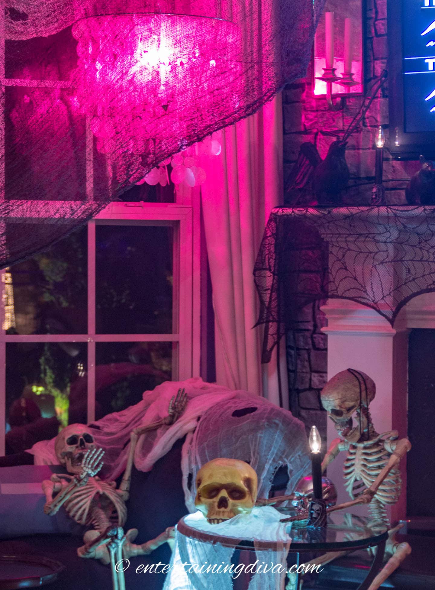 Skeletons sitting on a sofa covered in white creepy cloth in front of a window with black creepy cloth and a fireplace mantel with black lace