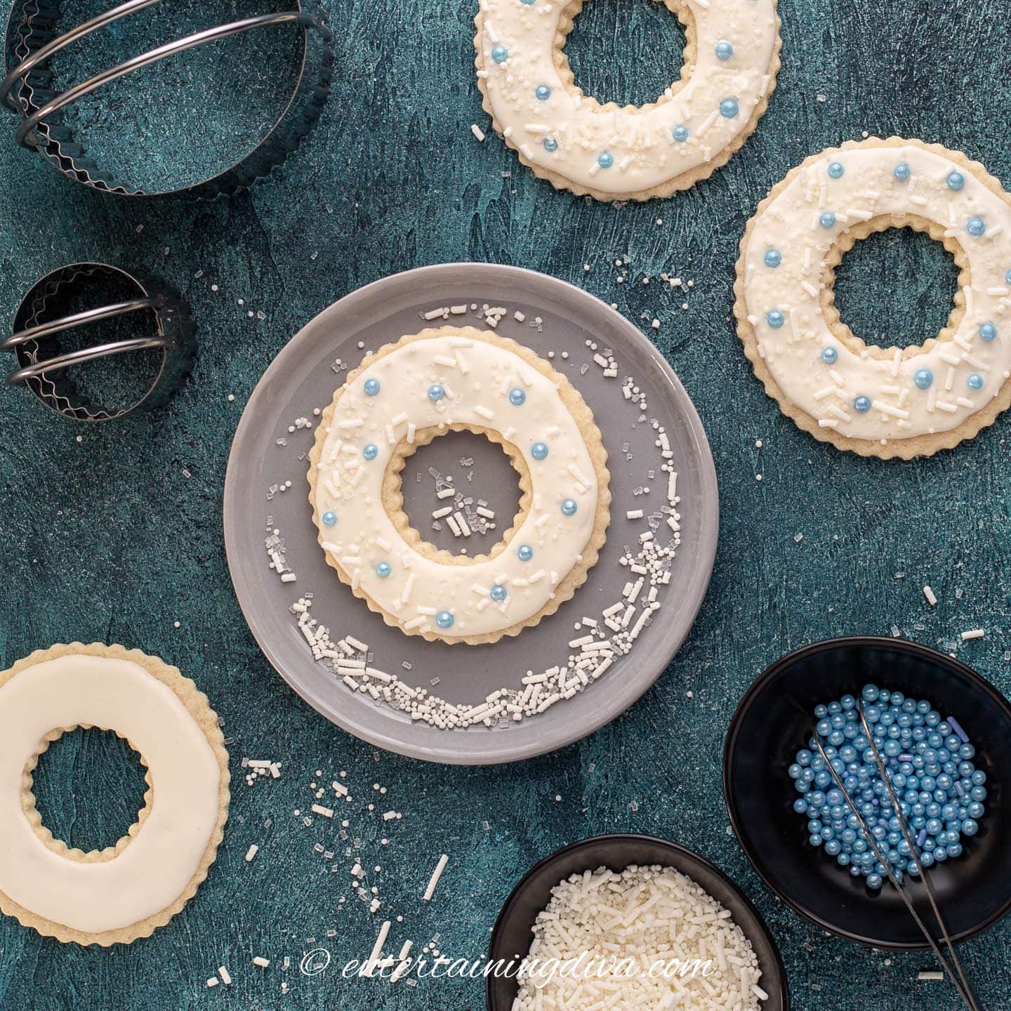 Wreath sugar cookie being decorated with white royal icing, white sprinkles and blue sugar balls