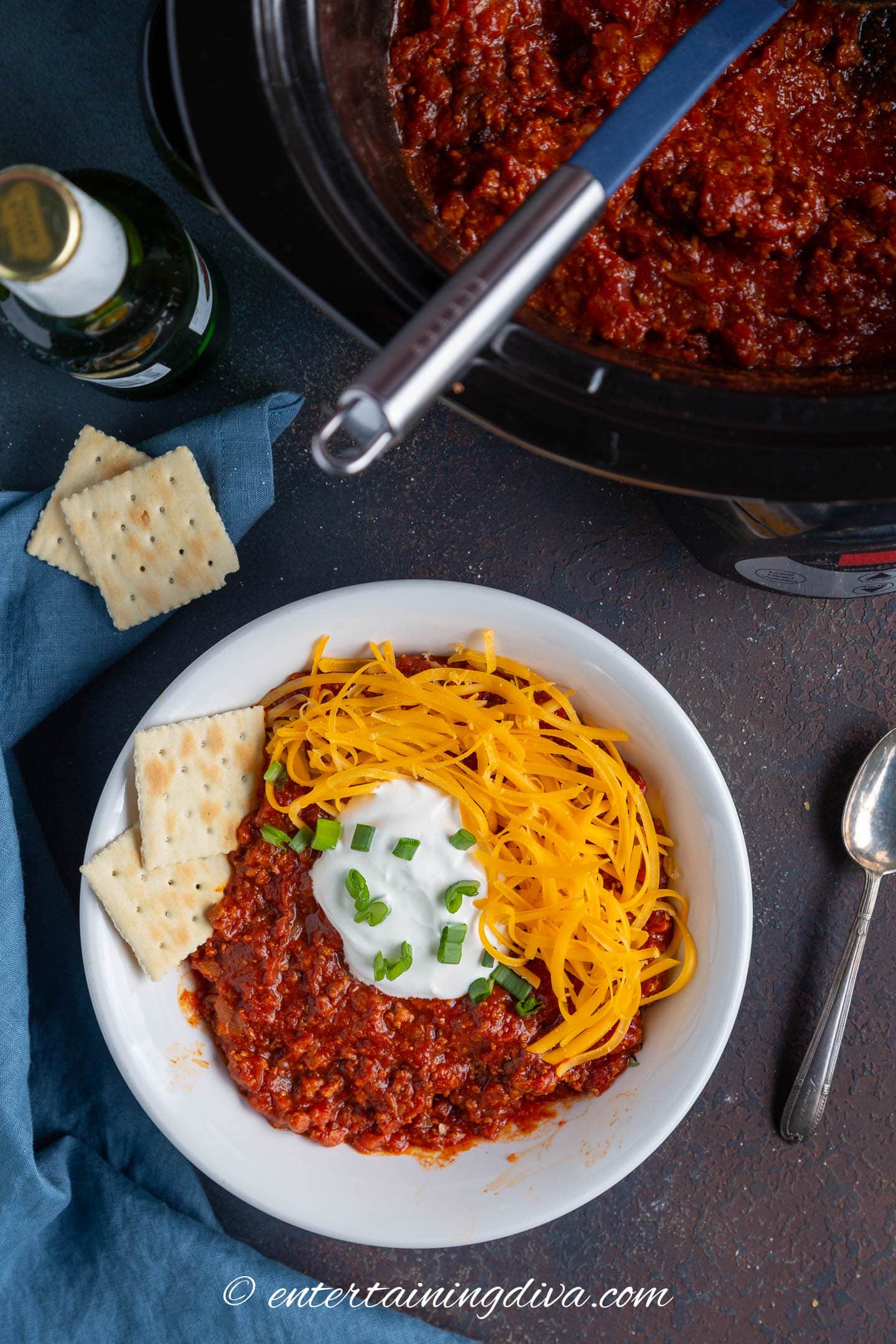 A bowl of crock pot no bean chili topped with sour cream, onions, grated cheese and crackers with a slow cooker in the background