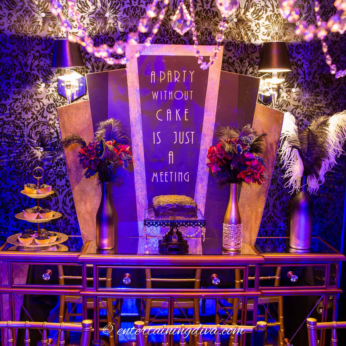 A Great Gatsby themed dessert table with cake, cupcakes and wine bottle vases with flowers and ostrich feathers